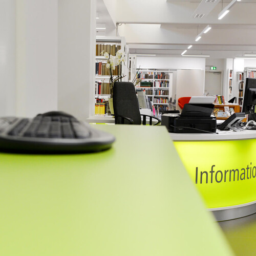 Infos zur Benutzung Linkerhand sieht man eine Tastatur im Vordergrund, rechterhand im Hintergrund sieht man die Infotheke der Bücherei Header_Infos_zur_Benutzung.jpg